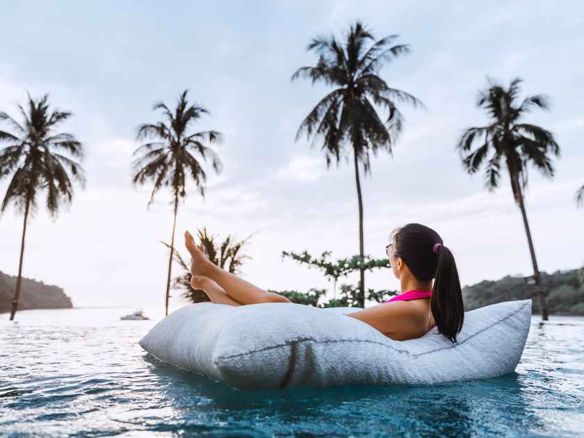 Summer travel vacation concept, Happy traveler asian woman with bikini relax on pool float in luxury infinity pool hotel resort with sea beach and palm tree background at sunset in Koh Kood, Thailand