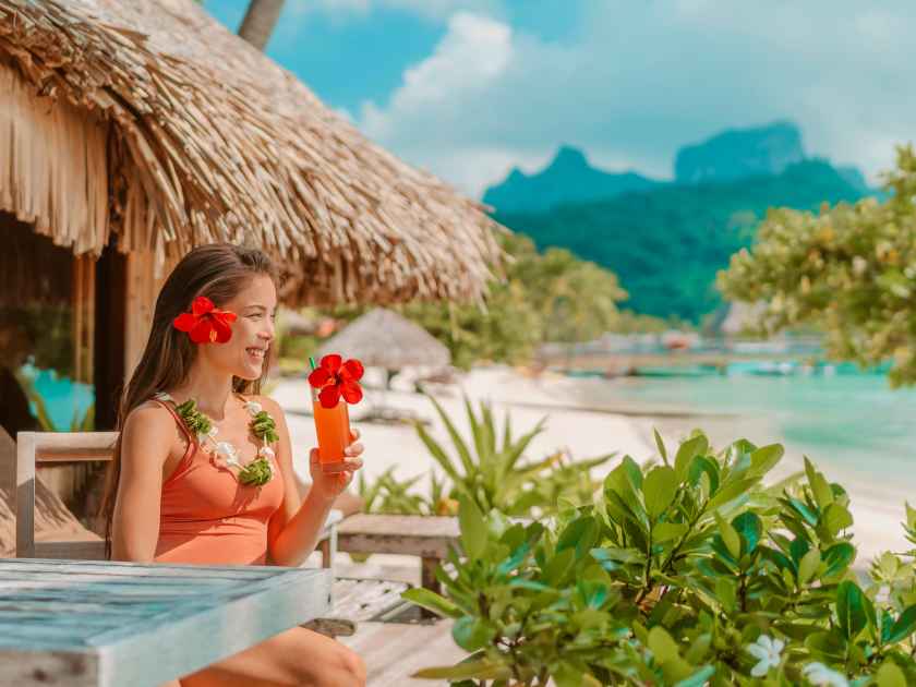 Breakfast at luxury hotel room on beach. Asian woman drinking fruit juice morning on summer vacation travel in Bora Bora island, Tahiti French Polynesia landscape. Happy tourist relaxing on holiday.
