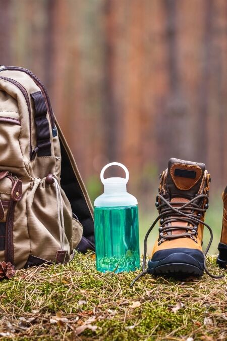 Hiking and camping equipment in forest. Backpack, water bottle and leather ankle boots. Panoramic view with copy space