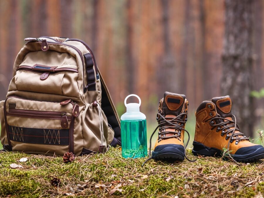 Hiking and camping equipment in forest. Backpack, water bottle and leather ankle boots. Panoramic view with copy space