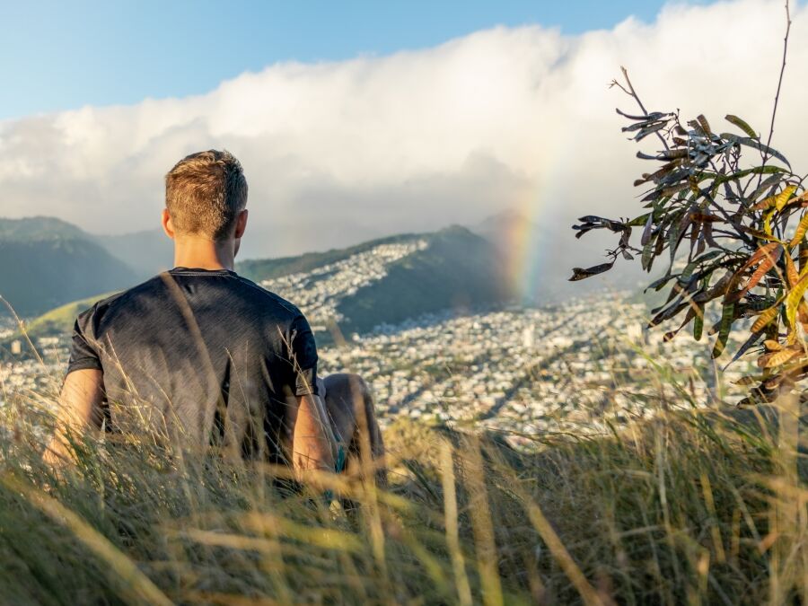 Young male hiker sitting on the summit of Diamond Head Crater in Honolulu on the Island of Oahu, Hawaii shortly before sunset, looking at a rainbow over the city and mountain range behind Honolulu.