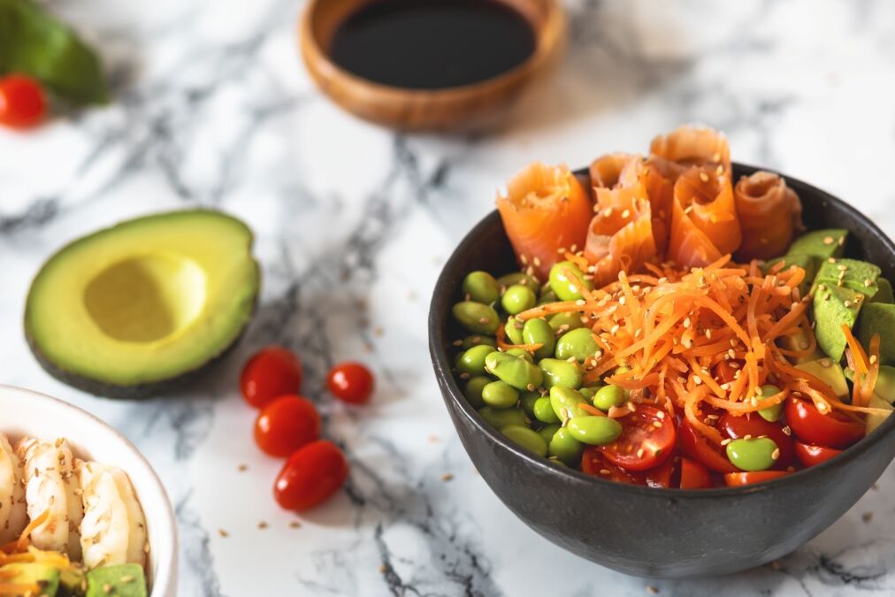 Nutritious poke bowl with avocado, edamame, cherry tomatoes, shredded carrots, shrimps and salmon on a marble background. Healthy food concept