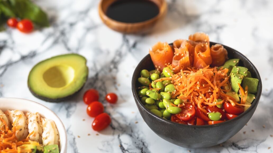Nutritious poke bowl with avocado, edamame, cherry tomatoes, shredded carrots, shrimps and salmon on a marble background. Healthy food concept