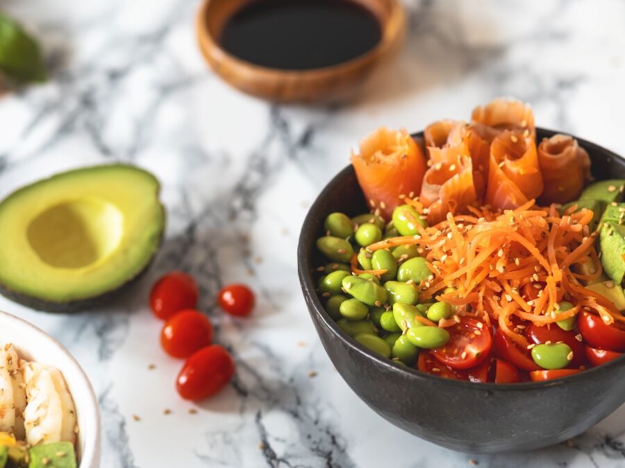 Nutritious poke bowl with avocado, edamame, cherry tomatoes, shredded carrots, shrimps and salmon on a marble background. Healthy food concept