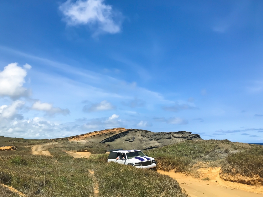 big cliff and dirt road against blue sky. landscape views of Papakōlea Green Sand Beach,Naalehu, Hawaii,United States on bright sunny days. White suv cars overcoming sand barrier. off road. copy space