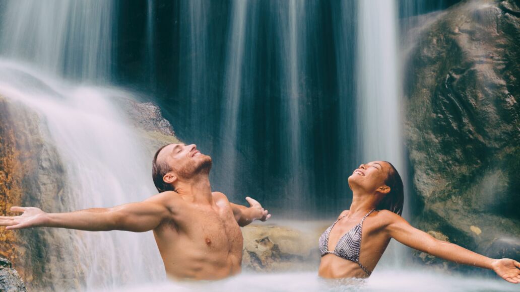 Nature,Couple,Relaxing,Under,Waterfall,With,Open,Arms,In,Freedom