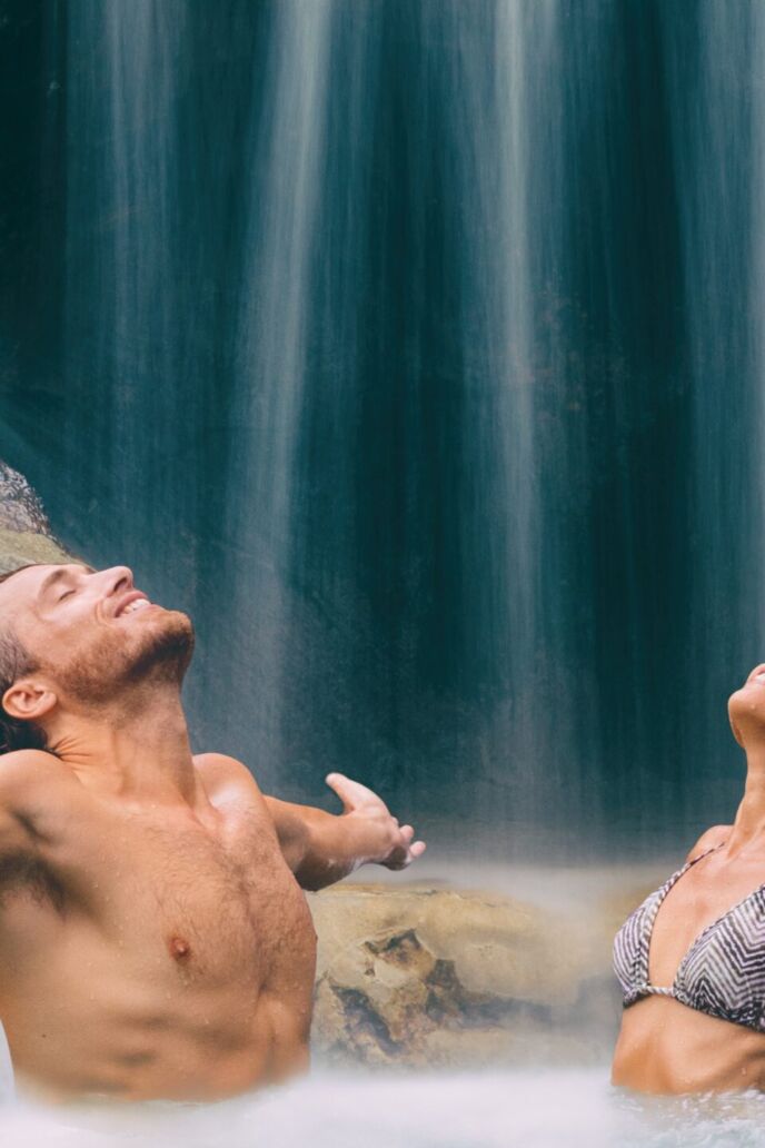 Nature,Couple,Relaxing,Under,Waterfall,With,Open,Arms,In,Freedom