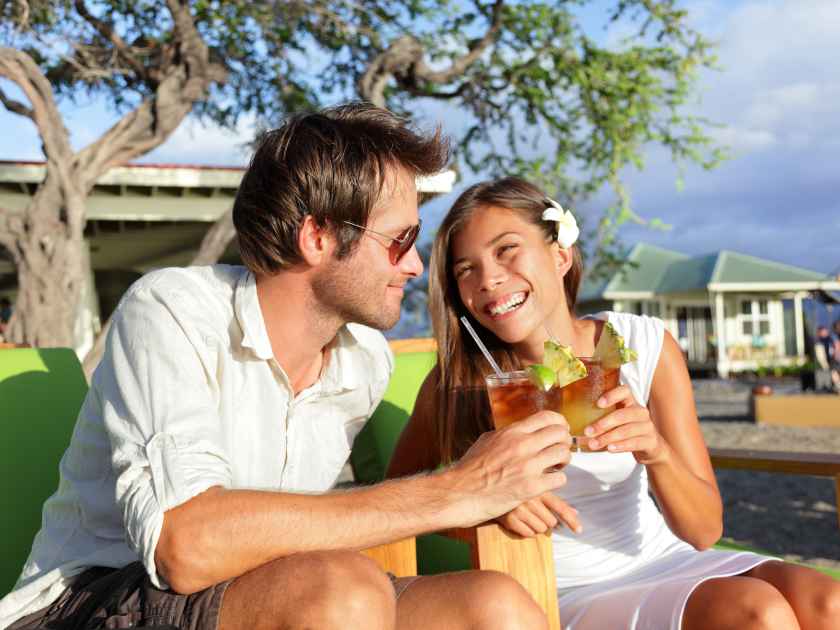 Couple drinking alcohol Mai Tai cocktails at Hawaii beach bar club at sunset. Beautiful asian girl flirting and enjoying alcoholic drinks outside on terrace with caucasian male.