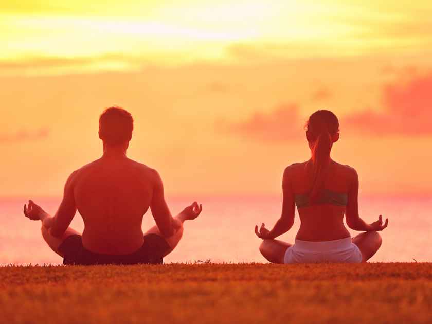 Meditation yoga couple meditating at serene beach sunset. Girl and man relaxing in lotus pose in calm zen moment in the ocean during yoga holiday class at resort retreat.