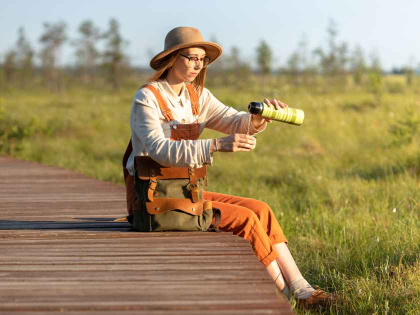 Woman botanist with backpack sitting on wooden path through peat bog swamp in wildlife national park. Naturalist resting on boardwalk, drinking tea, enjoying the moment at sunset. Ecotourism