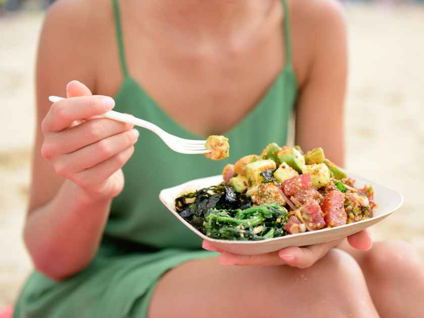 Poke bowl salad plate. A traditional local Hawaii food dish with raw marinated ahi yellowfin tuna fish. Woman sitting on beach eating lunch.