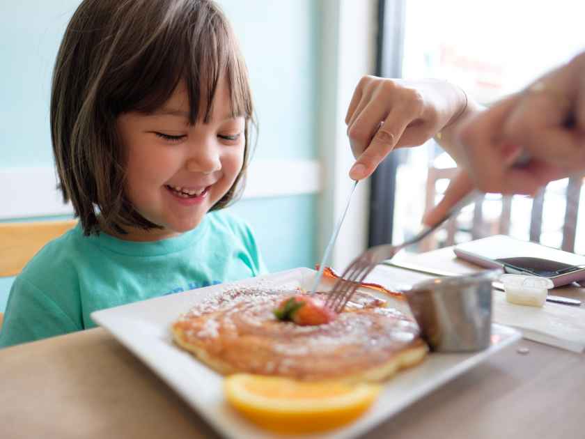 Little girl enjoying delicious pancakes with maple syrup and a strawberry for breakfast on a sunny summer day in Hawaii. Mother gives a helping hand.