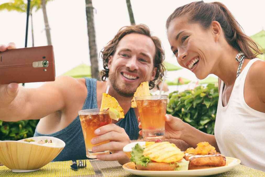 Selfie photo couple taking picture with phone at Hawaii beach bar on summer vacation. Happy Asian woman and man toasting mai tai drinks at bar having fun. Friends on travel holidays.