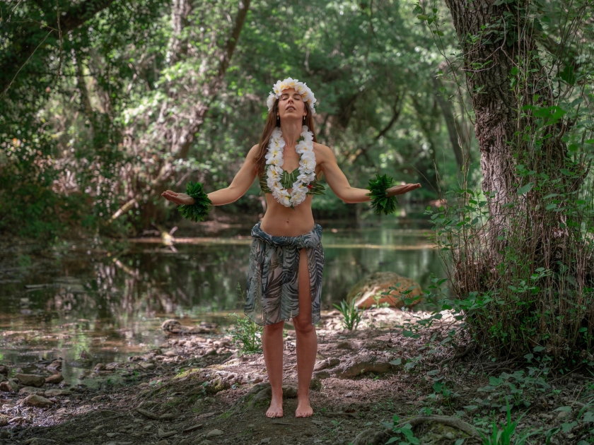 Happy young woman projecting positive energy and well-being in the middle of the forest wearing tropical hula dancer outfit. Miss Tropical wears a crown and necklace of white flowers.