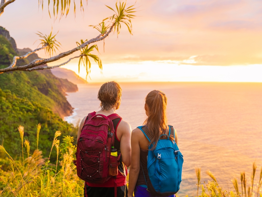 Hawaii hiking hikers hiking on Kalalau trail watching sunset from Na Pali Coast. Tourists couple with backpacks walking outdoor in Kauai island. Summer travel adventure active lifestyle.