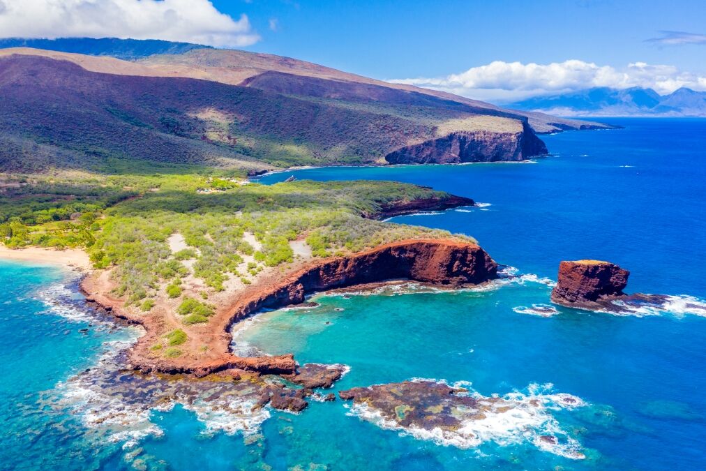 Aerial view of Lanai, Hawaii featuring Hulopo'e Bay and beach, Sweetheart Rock (Pu'u Pehe), Shark's Bay, and the mountains of Maui in the background.