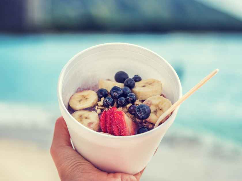 Acai bowl food selfie picture. Closeup of healthy breakfast take-out on ocean background at hawaii beach. Berries and fresh fruits outdoors for a weight loss diet. Square crop for social media.