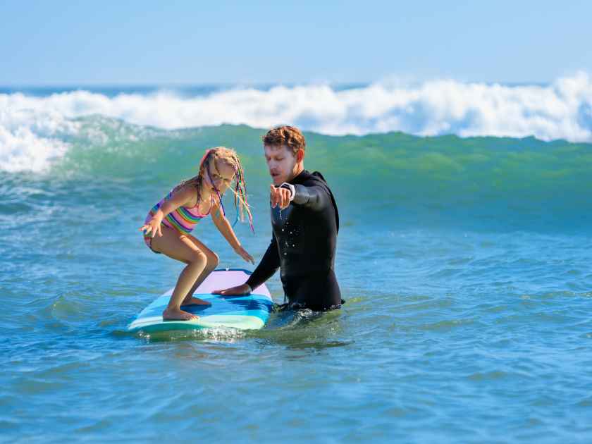 Little surf girl - young surfer learn to ride on surfboard with instructor at surfing school. Active family lifestyle, kids water sport lessons, swimming activity in summer camp. Vacation with child.