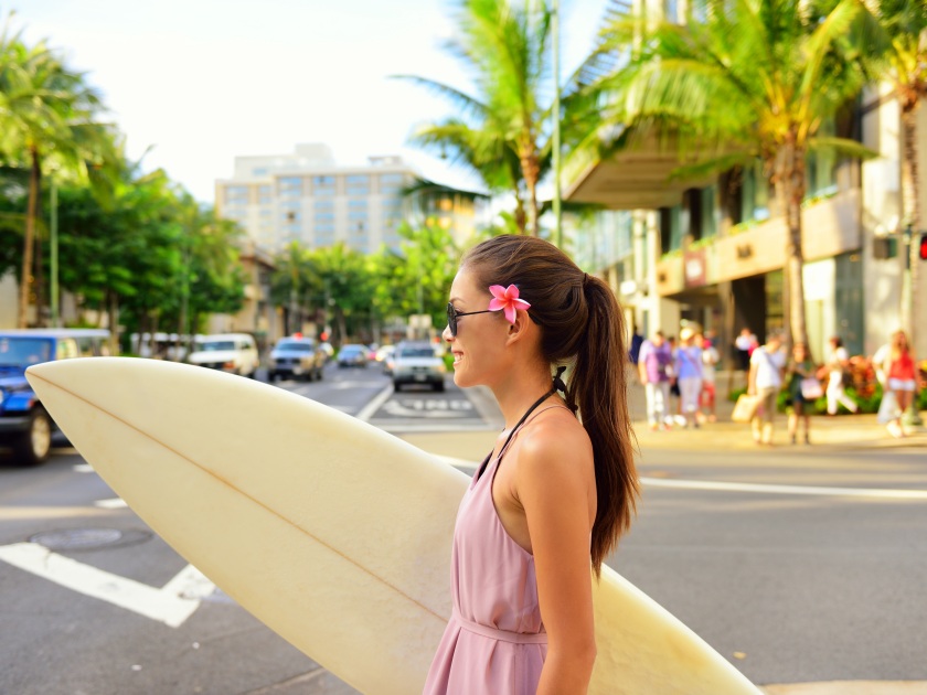 Surfer woman walking in city with surfboard to go surfing. Urban Hawaiian surf concept. Asian girl holding surf board crossing street to go to the beach. Waikiki, Honolulu city, Oahu, Hawaii, USA.
