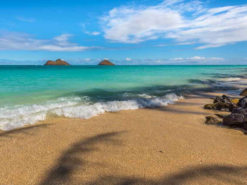 View of Na Mokulea (Mokulea Islands) from the shores of Lanikai Beach, in Kailua, Hawaii, Oahu - see the palm tree leaf shadows on the sand, as calm waves roll in on a sunny afternoon