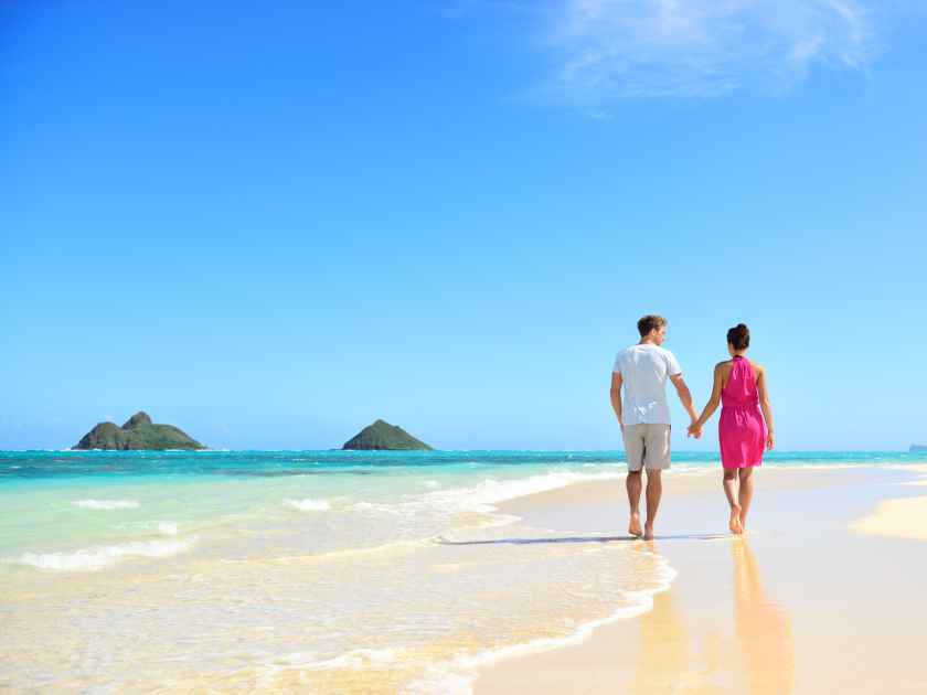 Beach honeymoon couple holding hands walking on white sand beach. Newlyweds happy in love relaxing on summer holidays on Lanikai beach, Oahu, Hawaii, USA with Mokulua Islands. Travel vacation concept.