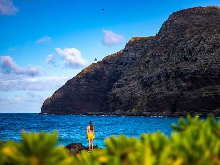 beautiful girl stands on the rocks in makapuu beach park admiring the sunset over makapu'u lighthouse on oahu, hawaiian islands; holiday in hawaii