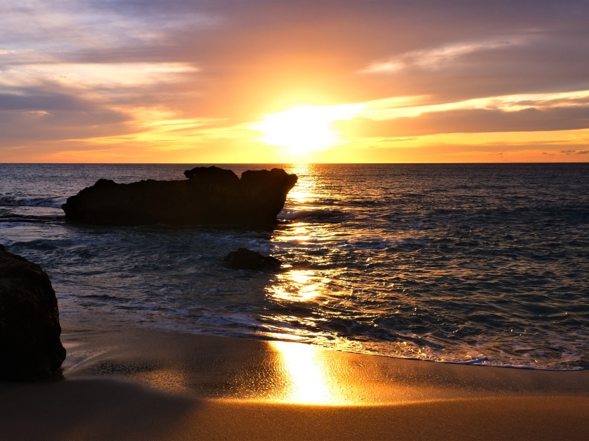 Dramatic gold orange sunset softening to lavender and blues by the end. Makua Beach, Oahu, HI. Large lava rock off shore, waves crashing over it and rolling into shore. Sunset reflecting on sand.