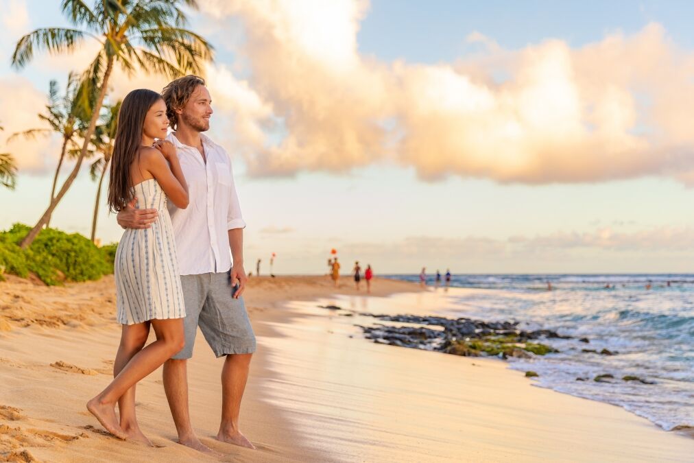 Couple on romantic sunset beach walk relaxing during Hawaii summer travel vacation on Kauai island, USA. Asian woman in white dress, Man in linen shirt.