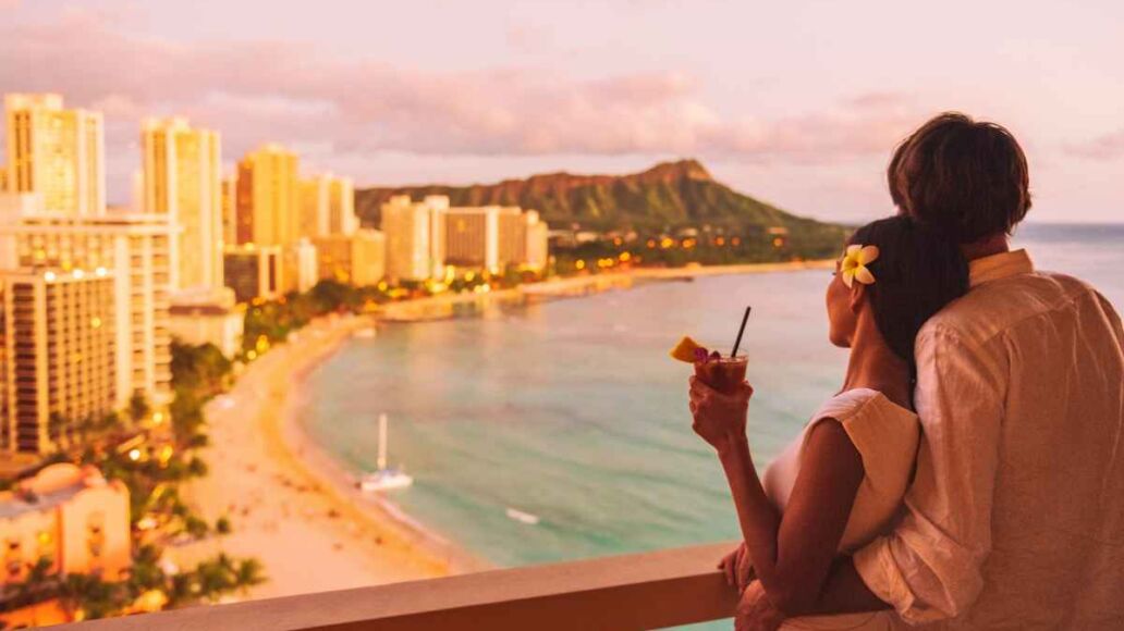 Hawaii luau vacation Mai Tai drink cocktail couple tourists at Honolulu hawaiian resort hotel. Happy man and woman relaxing at sunset view of Diamond Head, Oahu island travel.