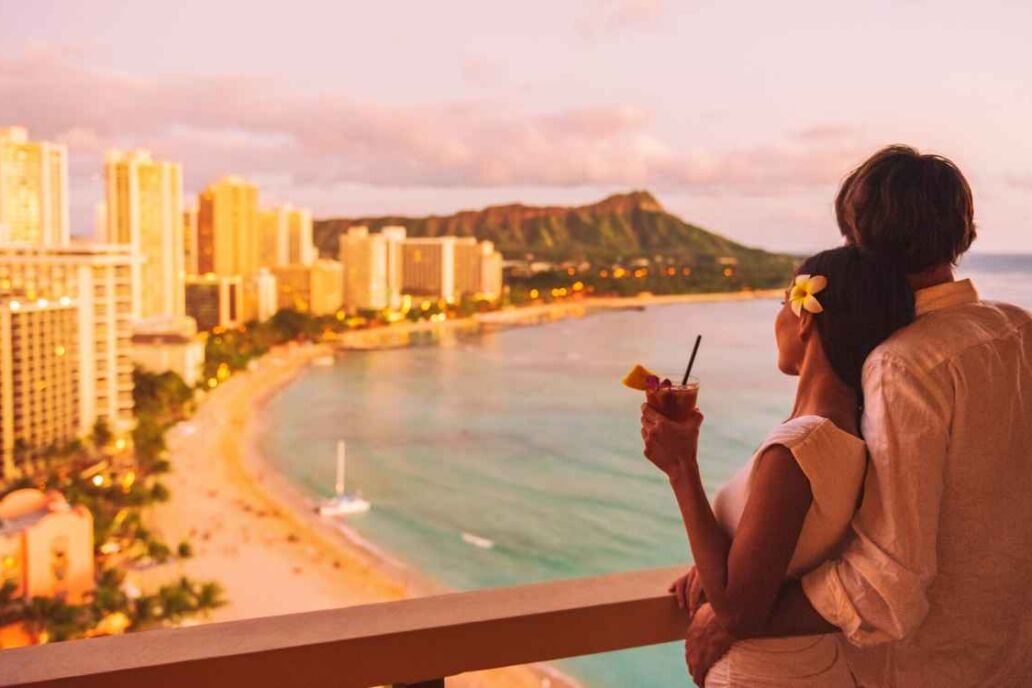 Hawaii luau vacation Mai Tai drink cocktail couple tourists at Honolulu hawaiian resort hotel. Happy man and woman relaxing at sunset view of Diamond Head, Oahu island travel.