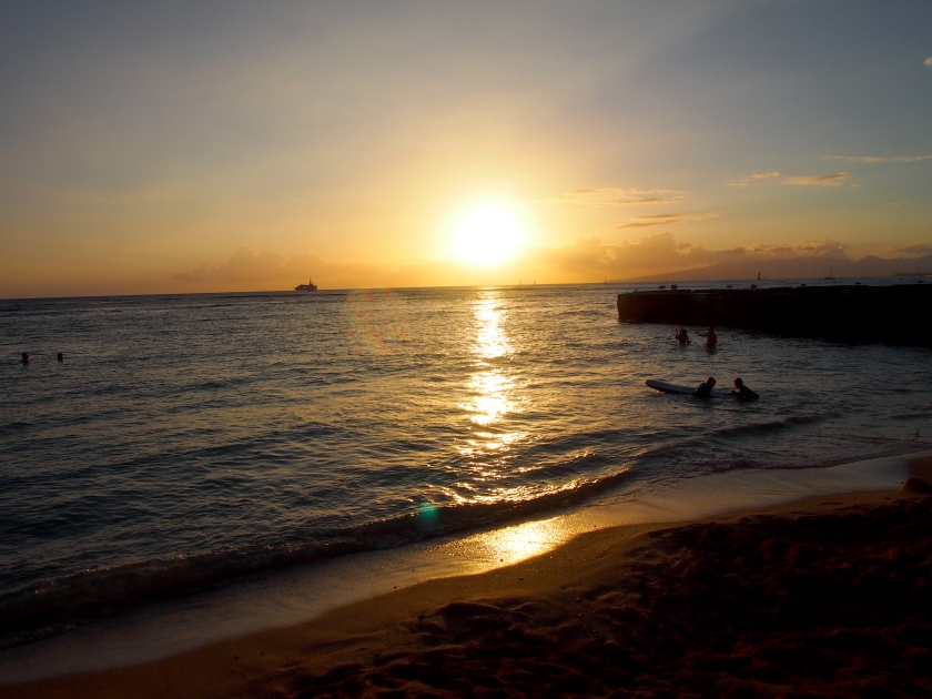 Sunset dropping behind the ocean on Kaimana Beach with boats on the water on Oahu, Hawaii.