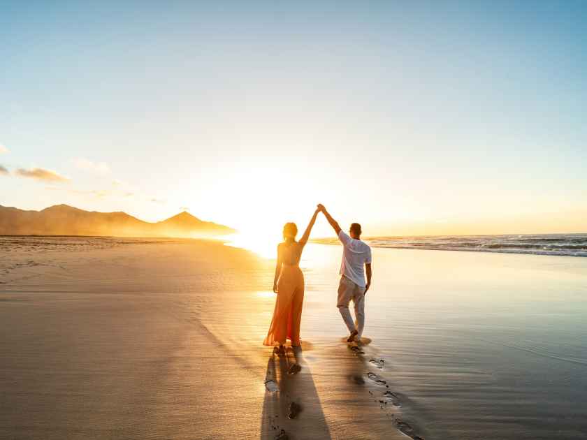Lovely, romantic couple walking on sunset beach, enjoying evening light, relaxing on tropical summer vacation. Honeymoon. Love. Back view. Woman wearing orange maxi dress.
