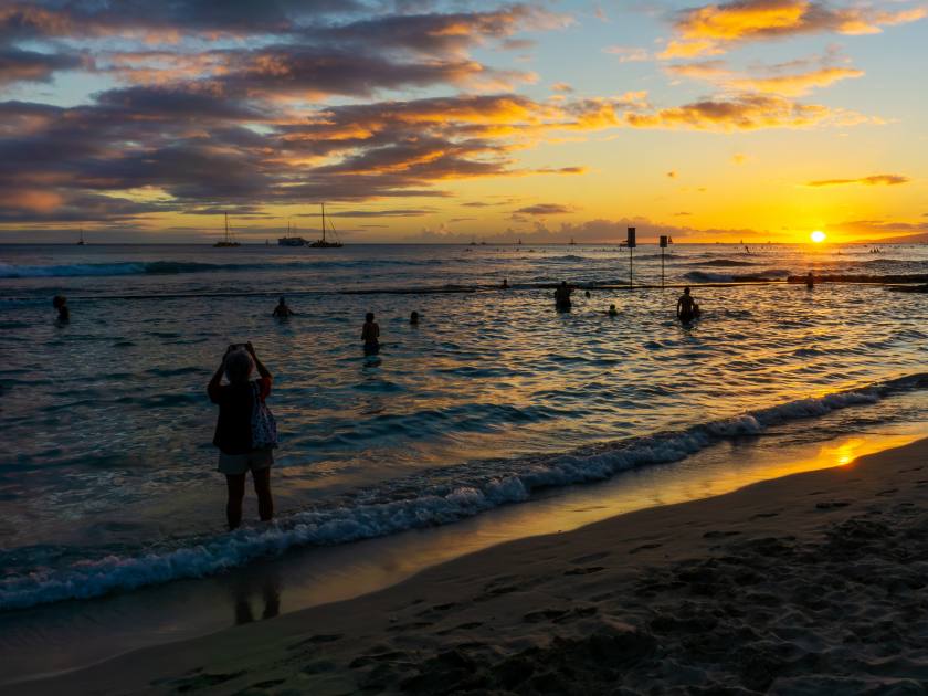 Golden sunset on horizon over Waikiki beach in Oahu, Hawaii, with silhouette of unidentifiable visitors swimming and on the beach.