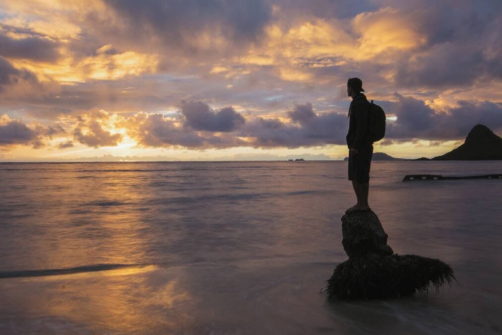 A person standing on a coconut tree stump in the shore line of Oahu's windward coast during a golden sunrise. The island known as China Man's Hat in the background.