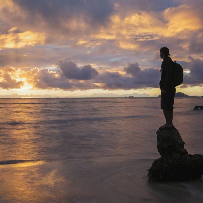 A person standing on a coconut tree stump in the shore line of Oahu's windward coast during a golden sunrise. The island known as China Man's Hat in the background.