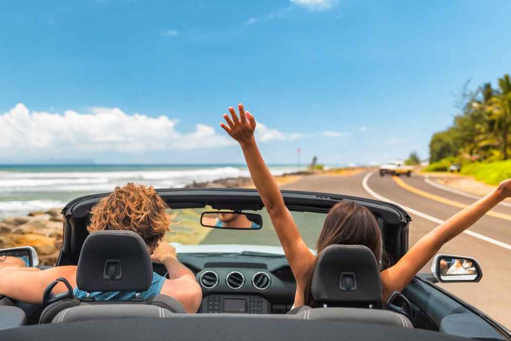 Road trip car holiday happy couple driving convertible car on summer travel Hawaii vacation. Woman with arms up having fun, young man driver