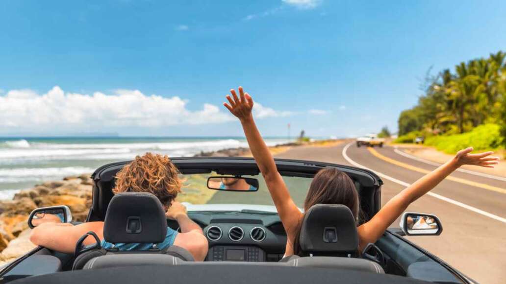 Road trip car holiday happy couple driving convertible car on summer travel Hawaii vacation. Woman with arms up having fun, young man driver