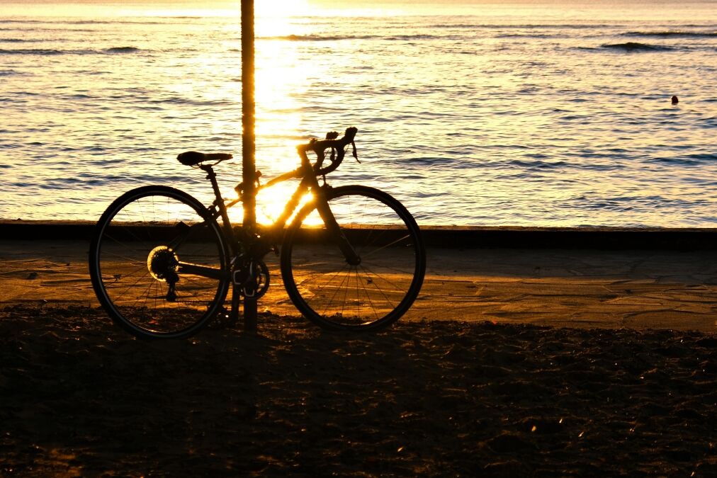 Golden sunset at Waikiki Beach with sailboats on the horizon. Silhouettes of hau tree, bicycle, palm trees and people. People silhouetted are watching sunset. Sky from light blue to yellow/orange.
