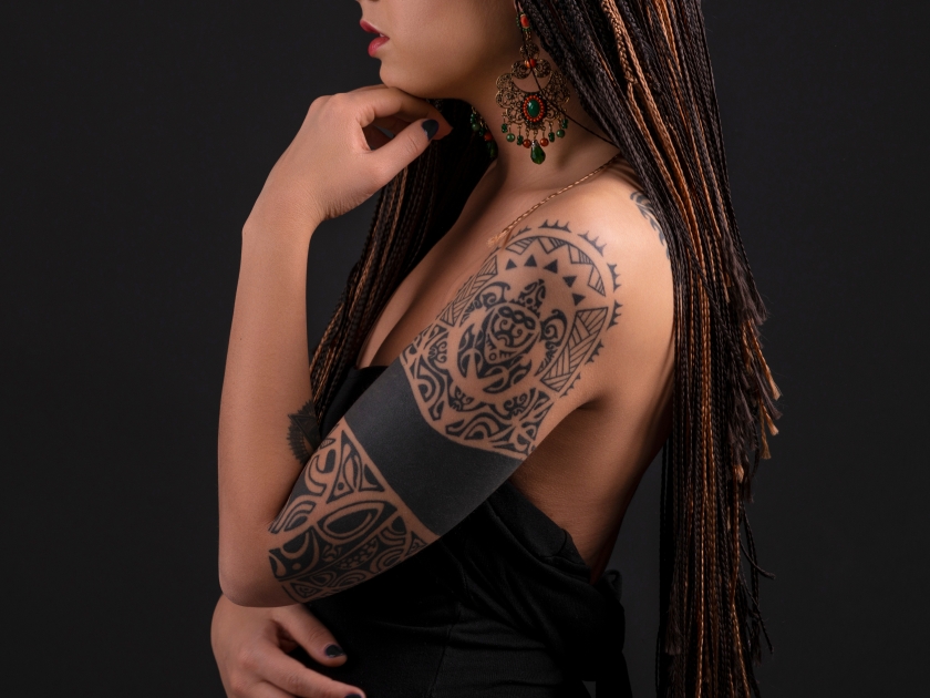 Half length portrait of a young woman with a polynesian tattoo on her arms and with african pigtails against dark background with space in the studio