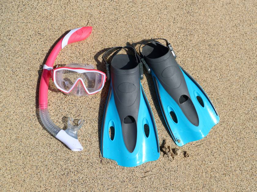 Beach vacation fun snorkel equipment with ocean waves splashing water. Scuba diving and snorkelling. Blue Flippers, pink mask, snorkel on sandy texture background. Objects lying on sand.