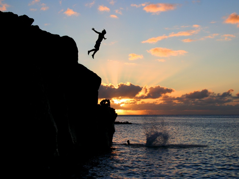 Boy jumps off a cliff into the ocean at Waimea Bay in Hawaii at sunset.